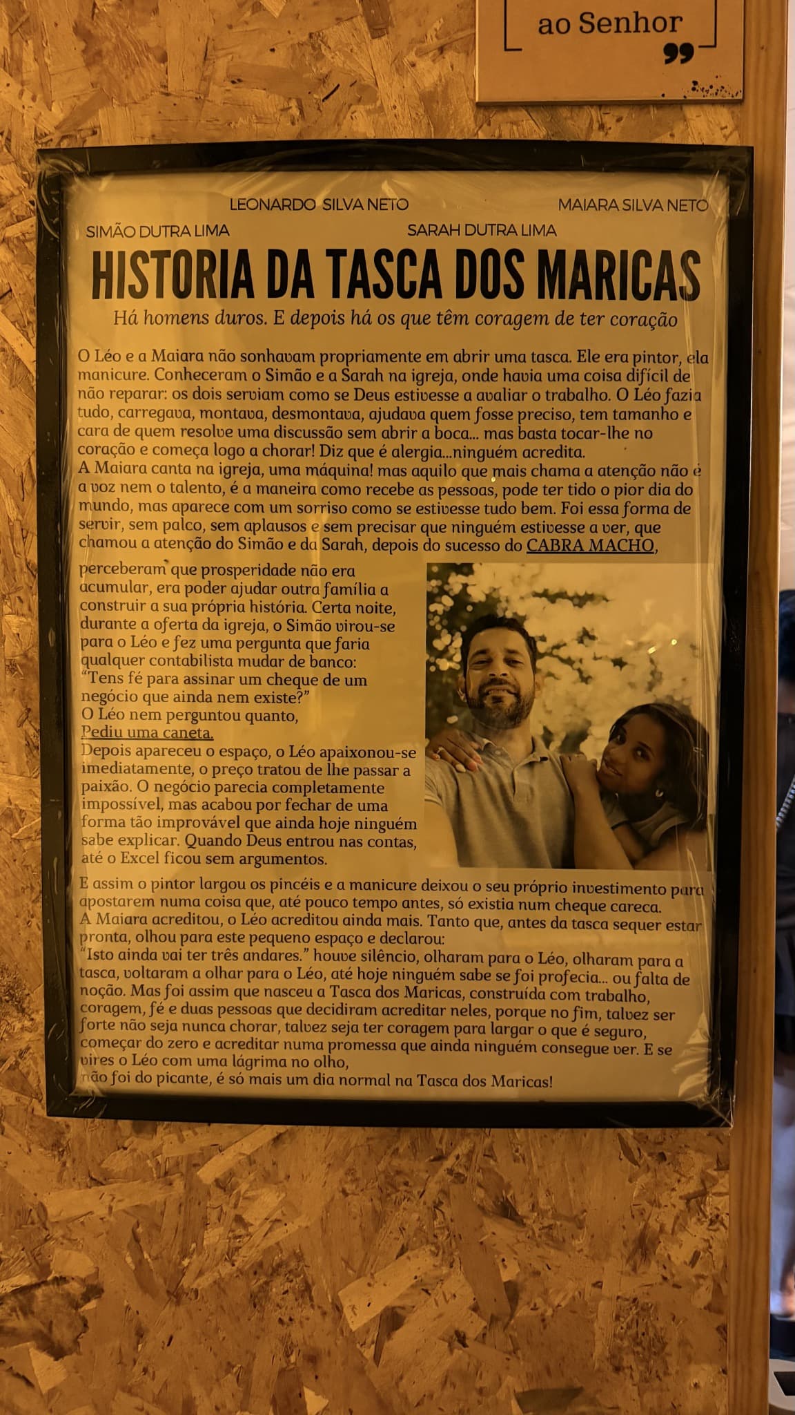 Quadro emoldurado com o texto “História da Tasca dos Maricas” e uma fotografia do Léo e da Maiara, pendurado numa parede de madeira.
