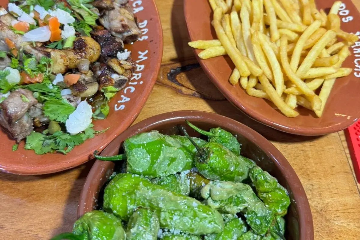 Mesa de madeira com pimentos padrón, batata frita e carne em pratos de barro da Tasca dos Maricas.